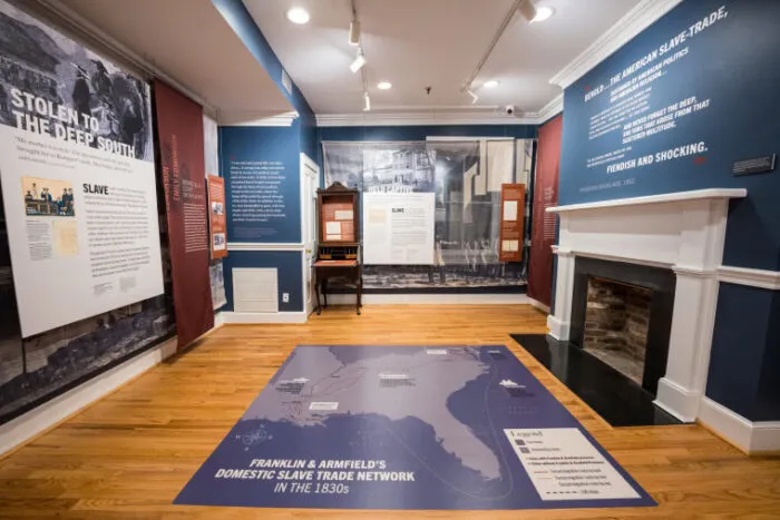 A museum exhibit room displays panels, a fireplace, and a map on the floor detailing the Franklin & Armfield slave trade network, with historical information and images about slavery in the U.S.