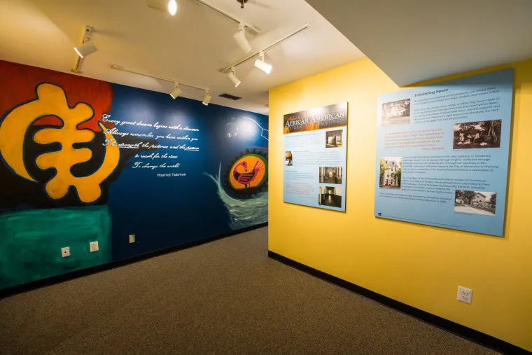 A small museum exhibit with colorful walls; one side features a large painted mural and a quote, while the other displays informational panels about African American history. Track lights illuminate the space.