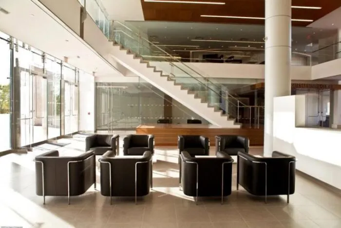 Modern office lobby with black leather chairs arranged in two rows, glass doors on the left, a staircase with glass railing in the background, and ample natural light filling the space.