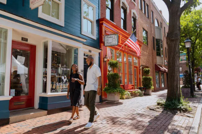 Two people walk and talk along a brick sidewalk lined with colorful storefronts and an American flag on a sunny day. The street has potted plants, large windows, and trees providing shade.