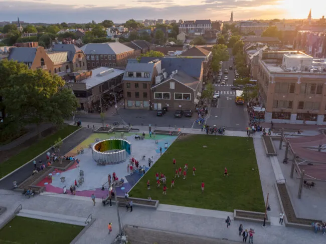 Aerial view of a city park at sunset, with people gathered around a colorful circular art installation, kids playing on the grass, and historic brick buildings in the background.