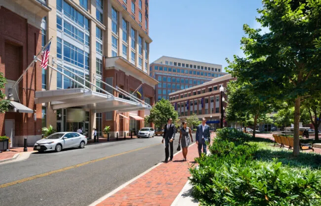 Three people in business attire walk on a brick sidewalk beside a modern office building with glass windows. Green trees line the street, and parked cars are visible. The weather is sunny and clear.