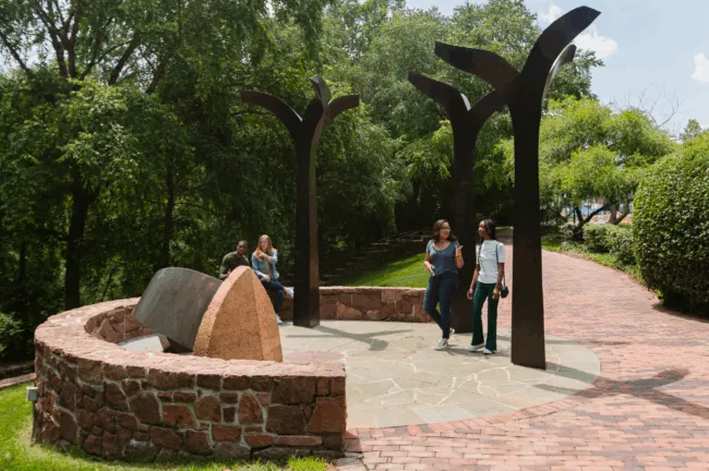 Four people interact at an outdoor memorial with metal tree-like sculptures and a large stone, surrounded by a low stone wall and trees, on a sunny day.