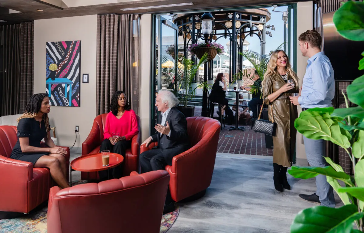 Five people socialize in a modern lounge. Three sit on red chairs talking, while two stand by a door leading outside, engaged in conversation. Sunlight and greenery are visible through the open door.