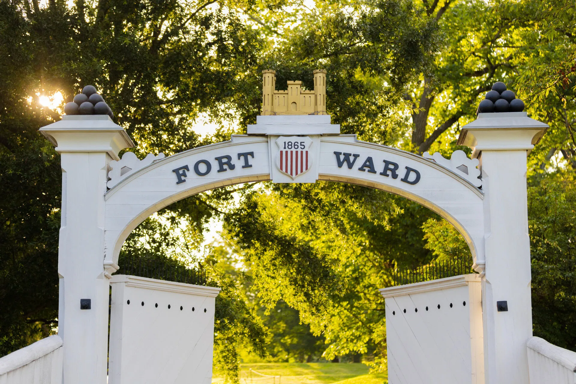 White arched entrance to Fort Ward with 1865 and a shield above, flanked by trees and greenery, sunlight streaming through leaves in the background.