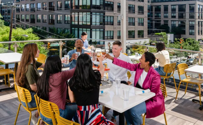 A diverse group of six people sit around a table on a rooftop patio, clinking glasses and smiling while enjoying food and drinks. City buildings and greenery are visible in the background.