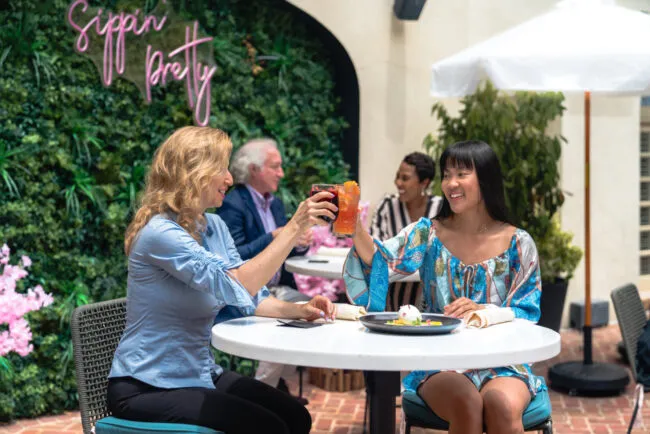 Two women sit at a round outdoor table toasting drinks and smiling. Behind them, a neon sign reads Sippin Pretty on a leafy wall. People sit at other tables, and a white umbrella shades the area.