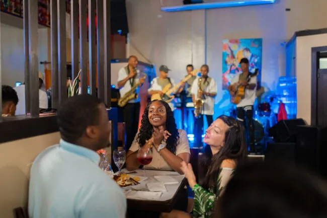 A live band performs in a colorful, lively restaurant as three people sit at a table in the foreground, smiling and enjoying food and drinks together. The atmosphere is cheerful and festive.