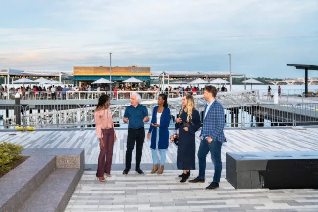 Five people stand and chat on a waterfront promenade, with a lively, modern restaurant and outdoor seating area in the background. The sky is clear and the scene is relaxed and social.