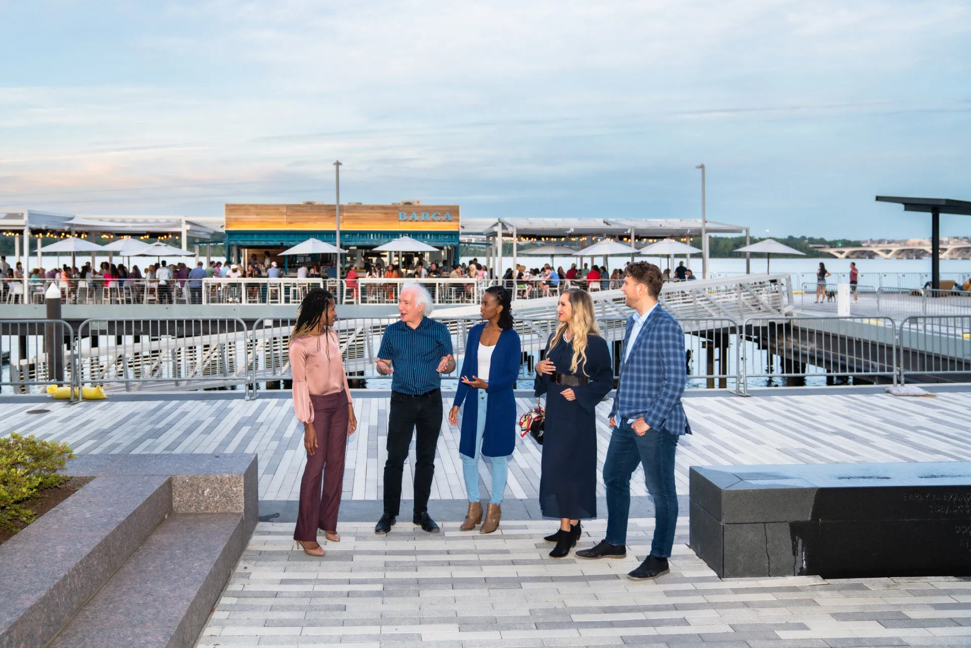 Five people stand and chat on a waterfront promenade, with a lively, modern restaurant and outdoor seating area in the background. The sky is clear and the scene is relaxed and social.