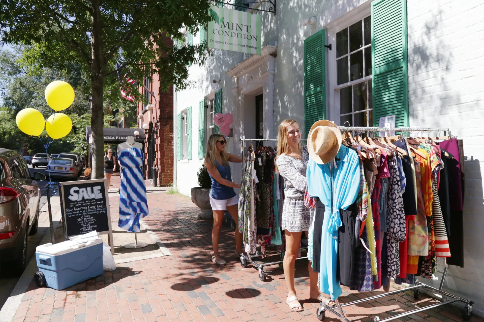 Two women browse colorful dresses on racks outside a boutique with green shutters. A SALE sign and yellow balloons are by the entrance on a sunny day, with shadows from nearby trees on the sidewalk.