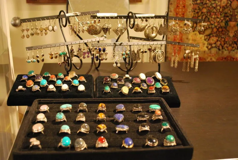 A display of colorful gemstone rings and various dangling earrings arranged on black trays and metal stands in a jewelry store. The background includes patterned fabric.