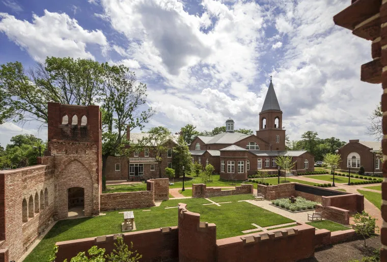 A scenic view of a historic brick courtyard with green lawns, surrounded by red brick buildings and a church with a steeple, under a partly cloudy sky.