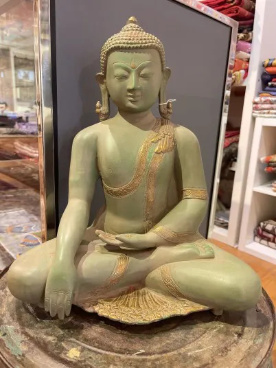 A green Buddha statue sits cross-legged on a pedestal, with one hand resting on the knee and the other in its lap. Shelves with colorful fabrics are visible in the background.