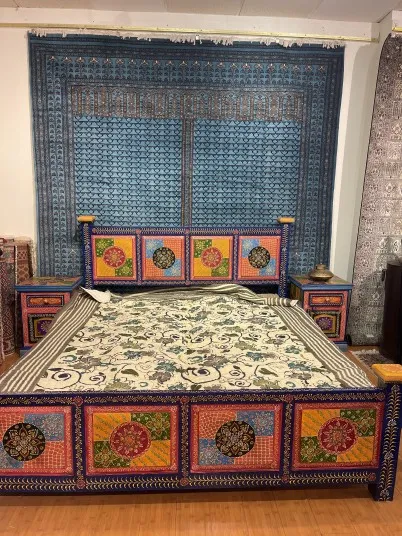 A colorful, hand-painted bed with matching side tables features intricate floral patterns in bright red, orange, yellow, and blue. A blue patterned tapestry hangs behind the bed, covering most of the wall.