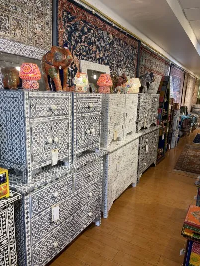 A row of intricately patterned white and gray dressers with inlaid designs is displayed in a store, along with mosaic lamps, elephant statues, and decorative rugs hanging on the walls.