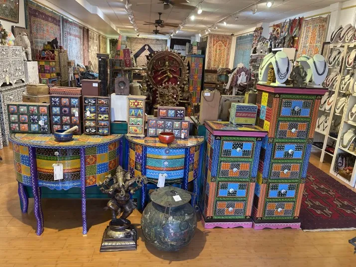 A colorful shop displays intricately painted wooden furniture, decorative boxes, statues, rugs, jewelry, and wall hangings. The vibrant patterns and diverse items create a lively, eclectic atmosphere.