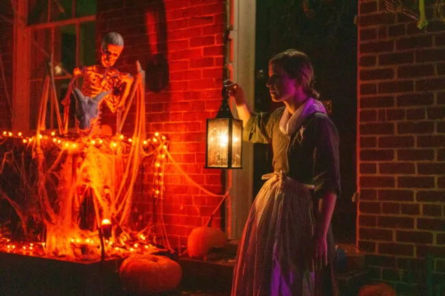 A woman in historical dress holds a lantern at night beside a brick house decorated with orange lights, cobwebs, pumpkins, and a blue skeleton for Halloween.