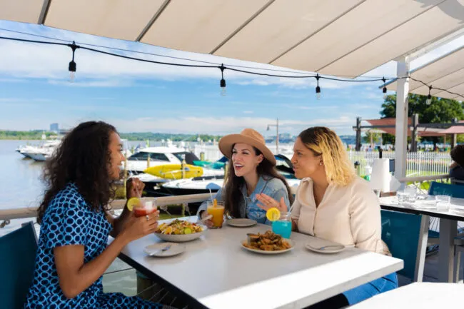 Three women sit at an outdoor restaurant table by the water, enjoying drinks and meals, smiling and talking. Boats are docked in the background under a sunny sky with a canopy overhead.