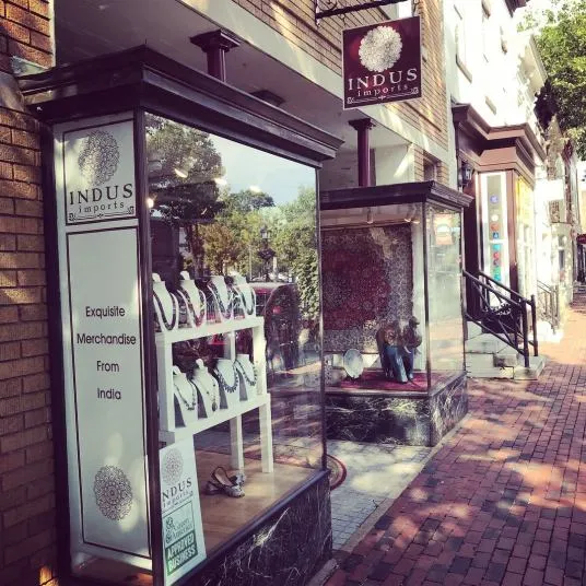 A storefront for Indus Imports displays jewelry in the window with a sign reading Exquisite Merchandise From India. The sidewalk is brick, and neighboring shops are visible along the street.