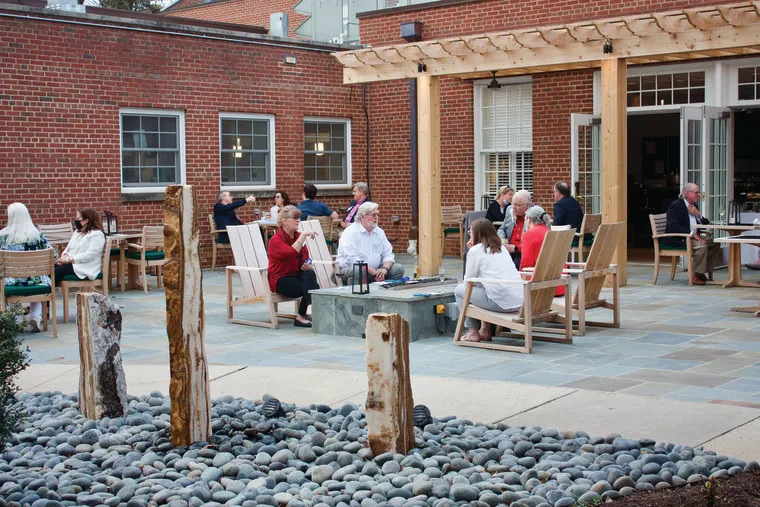 People sit and converse at outdoor tables on a stone patio surrounded by brick buildings, wooden beams, and a decorative area with smooth gray rocks and upright wooden posts in the foreground.