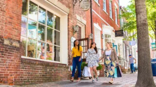 Three women walk on a brick sidewalk, carrying shopping bags and smiling, in front of brick buildings and shop windows on a sunny day. A man can be seen walking in the background.