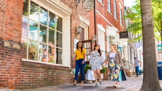 Three women walk on a brick sidewalk, carrying shopping bags and smiling, in front of brick buildings and shop windows on a sunny day. A man can be seen walking in the background.
