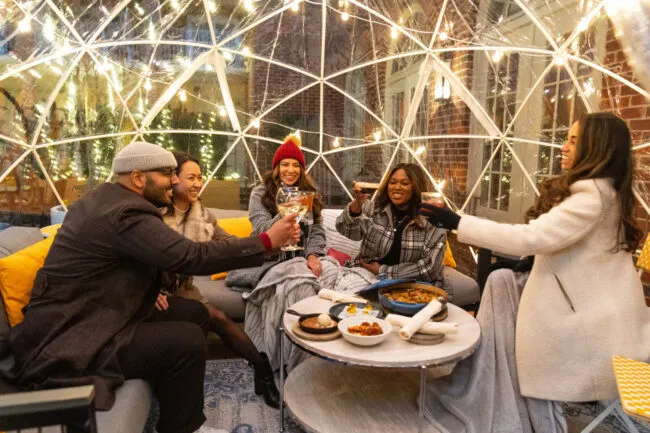 Five friends sit in a cozy outdoor igloo, clinking glasses and smiling around a table with food, under warm string lights on a chilly evening.
