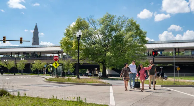 A group of five people cross a street at a crosswalk near a train station on a sunny day, with a large green tree and a historic-looking tower in the background.