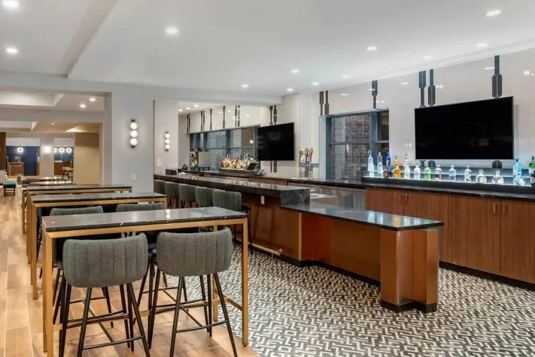 Modern bar area with tall tables and gray cushioned stools, a long wooden bar with black countertop, two large wall-mounted TVs, decorative lighting, and a mix of wood and patterned tile flooring. Bottles are displayed behind the bar.