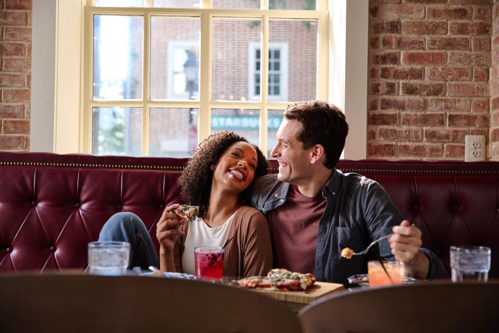 A couple sits in a red booth at Mia's Italian Kitchen smiling and enjoying pizza and cocktails.