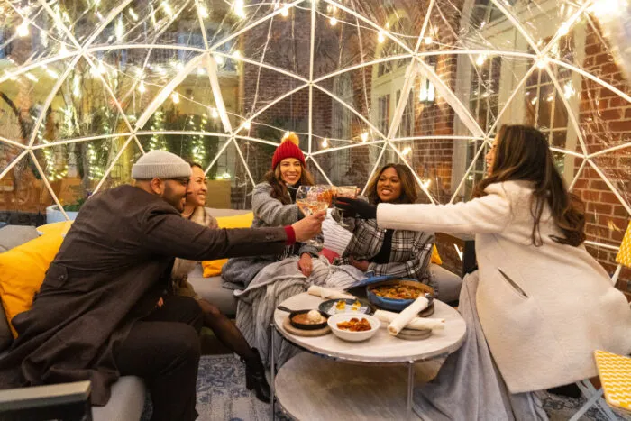 Four friends sit inside a clear dome decorated with string lights, bundled in coats and hats, smiling and clinking glasses over a table with snacks on a cozy outdoor patio.