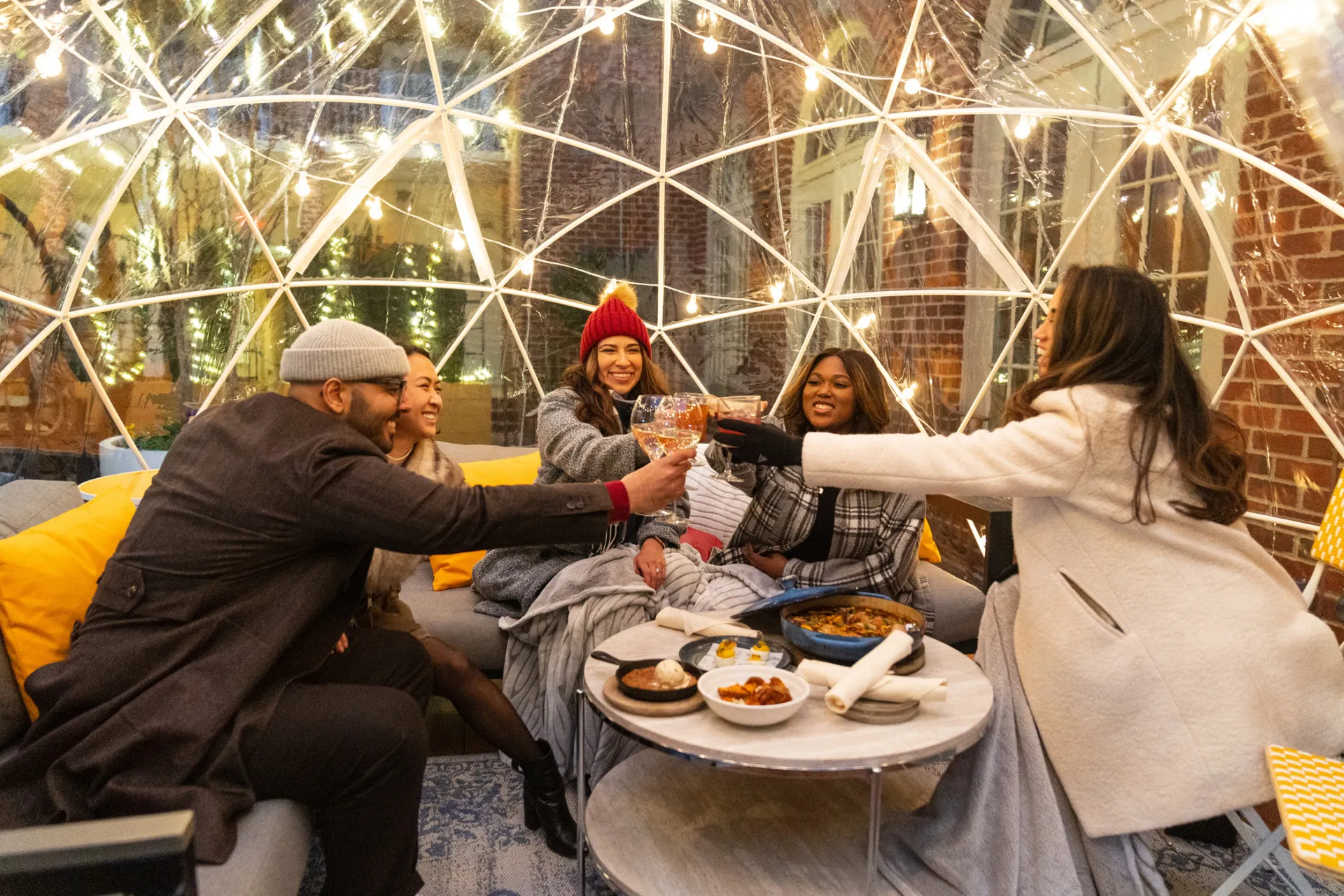 Four friends sit inside a clear dome decorated with string lights, bundled in coats and hats, smiling and clinking glasses over a table with snacks on a cozy outdoor patio.