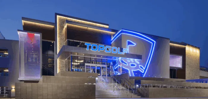 A modern Topgolf building at dusk, illuminated by vibrant blue and white neon lights, with large glass windows and prominent signage above the entrance.