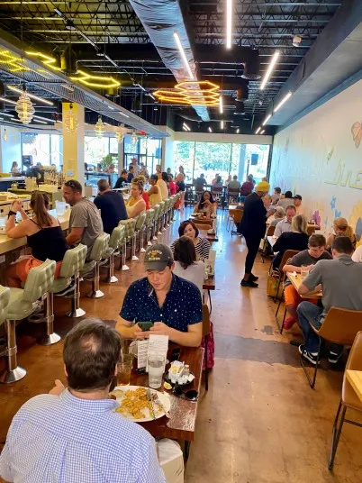 A busy, brightly lit restaurant with people dining at tables and a long counter. Diners are engaged in conversation, and staff move between tables. Large windows at the front let in natural light.