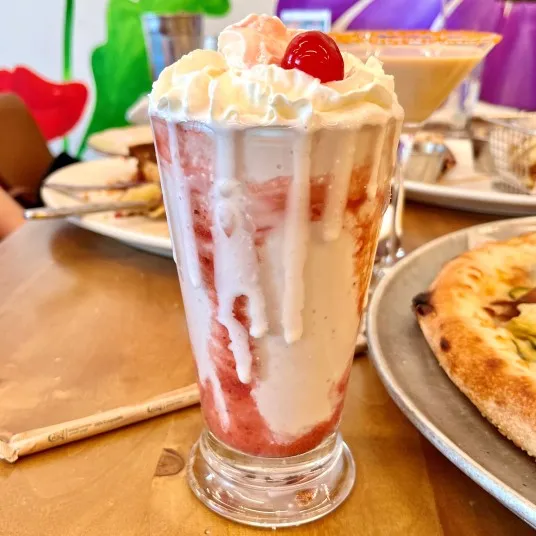 A tall glass of vanilla and strawberry milkshake topped with whipped cream and a cherry sits on a wooden table next to a pizza and other food items.