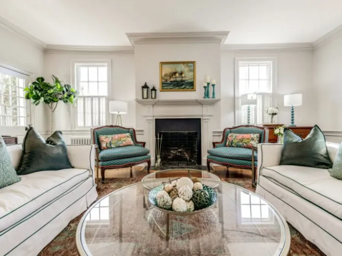 Bright living room with two white sofas facing each other, green accent pillows, a glass coffee table with decorative spheres, a fireplace with a ship painting above it, and large windows letting in natural light.