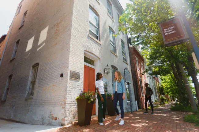 Two women stand and talk outside the Freedom House Museum, a historic brick building with a white door on a tree-lined sidewalk. A museum sign hangs from a post nearby. Another person walks further down the sidewalk.
