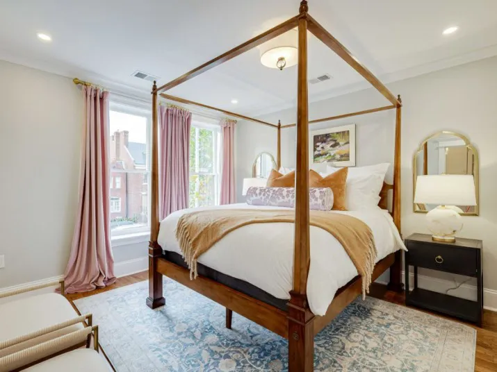 A bright bedroom with a four-poster bed, white linens, tan and mauve pillows, two mirrors, a nightstand with a lamp, and large windows with pink curtains letting in natural light.