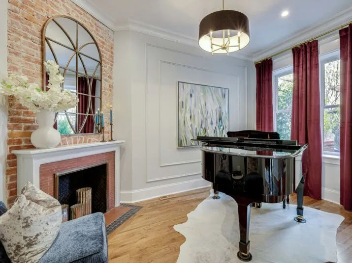 A stylish living room with a black grand piano on a white rug, red curtains, large windows, a brick fireplace with a round mirror, and a modern chandelier hanging from the ceiling.