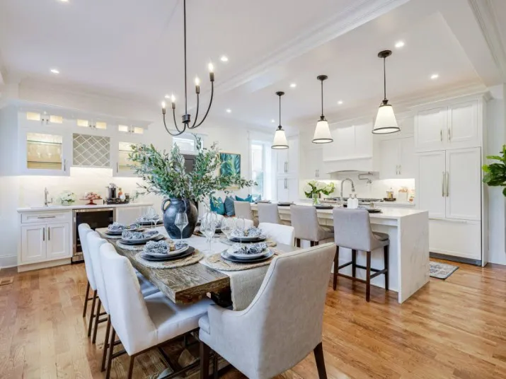Modern, spacious kitchen and dining area with white cabinetry, a large island with bar stools, elegant dining table set for six, hardwood floors, pendant and chandelier lighting, and decorative greenery.