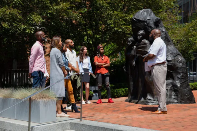 A group of people stands outdoors around a large bronze statue, listening to a man speaking. The scene is set on a brick path with greenery and buildings in the background.