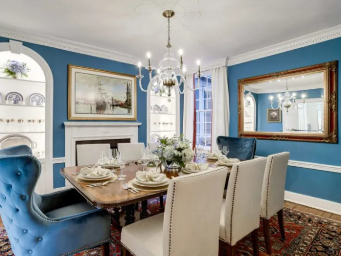 A formal dining room with a wooden table set for six, white and blue upholstered chairs, a chandelier, blue walls, a large mirror, framed ship artwork, and decorative shelves with plates.