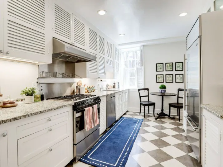 Bright, modern kitchen with white cabinets, stainless steel appliances, a blue rug on a checkered floor, and a small round table with two chairs by a window with natural light.