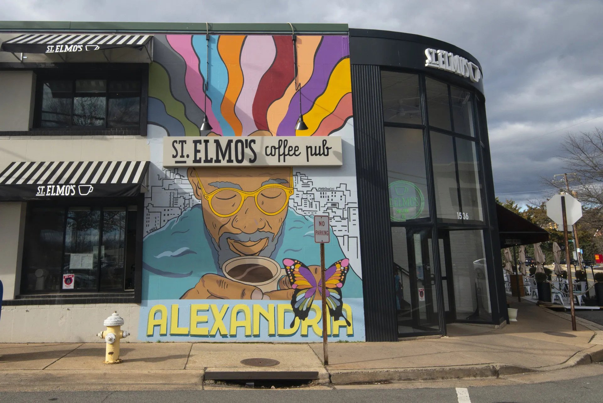 A mural on the side of St. Elmo’s Coffee Pub in Alexandria shows a colorful portrait of a man with sunglasses holding a coffee cup, with vibrant swirls above his head and a butterfly below.