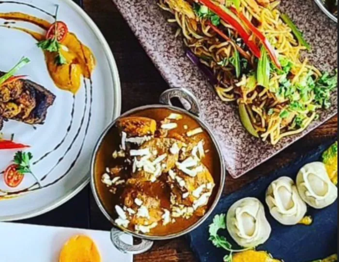 A top-down view of assorted Indian dishes, including curry in a metal bowl, vegetable noodles, dumplings, grilled meat, sauces, and garnishes on decorative plates.