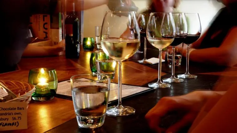 A group of people sitting around a wooden table with glasses of wine, water, menus, and lit green votive candles, creating a warm, cozy atmosphere. Some hands are visible as people interact.