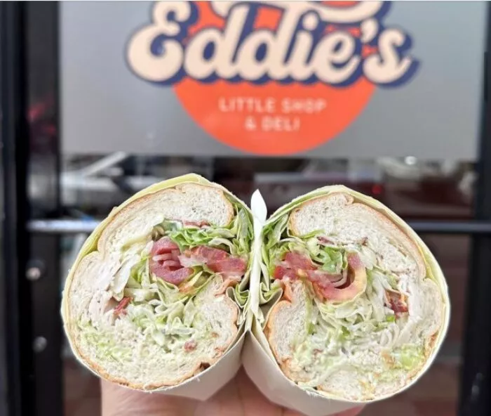 A hand holds a sandwich cut in half, filled with bacon, lettuce, tomato, and mayonnaise, in front of a glass door with the “Eddie’s Little Shop & Deli” logo.
