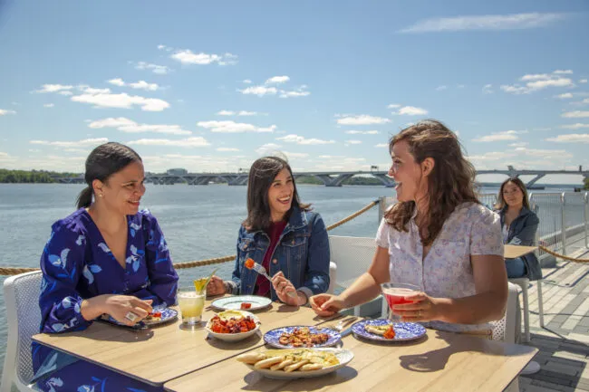 Three women sit at an outdoor restaurant table by the water, laughing and enjoying food and drinks on a sunny day. A bridge and a fourth person sitting alone are visible in the background.