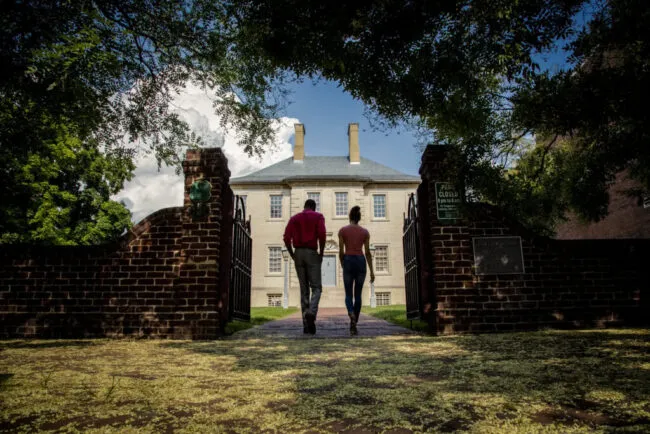 Two people walk hand-in-hand through an open brick gate toward a large historic building, framed by leafy trees and a blue sky with clouds above.